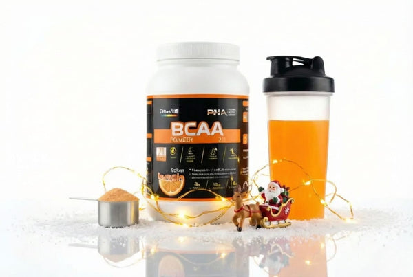 Embalagem de PNA BCAA Powder 2:1:1 sabor laranja ao lado de um shaker com bebida cor de laranja e colher doseadora, decorados com luzes de fada e um Pai Natal num trenó sobre neve artificial.
